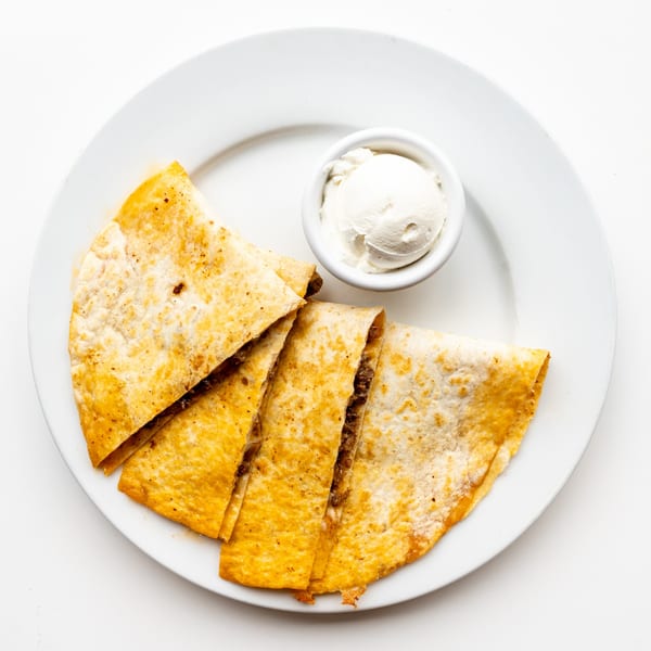 Four quesadilla slices filled with meat, served with a side of sour cream on a white plate.