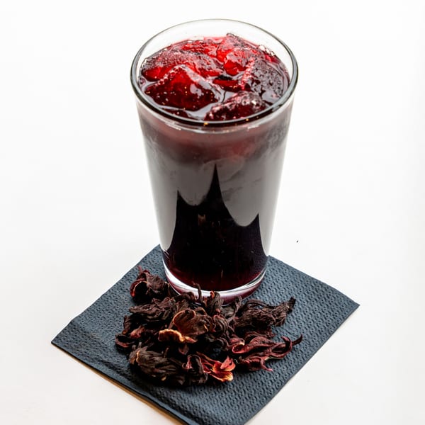 A glass of dark red hibiscus agua fresca with ice, next to dried hibiscus flowers on a napkin.