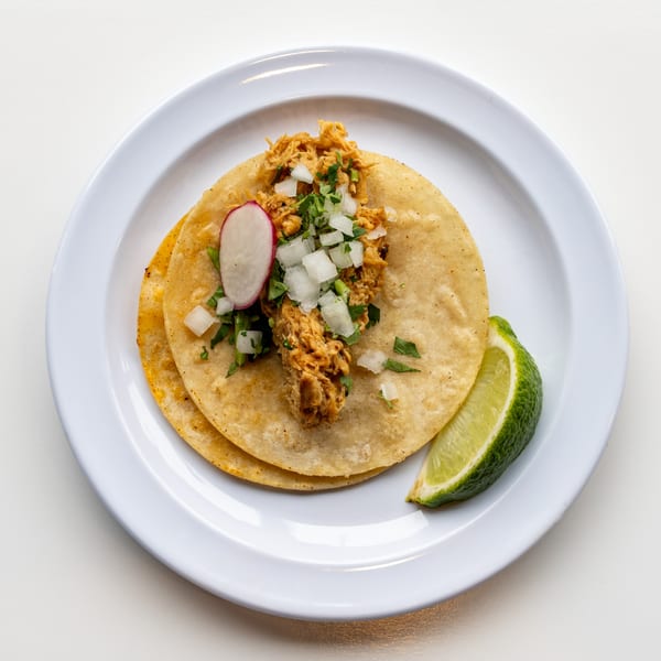 Two Casa De Avila Tacos with shredded chicken, onions, cilantro, radish, and lime on a white plate.
