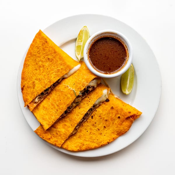 Four birria tacos with melted cheese and meat, served with a side of consommé and lime wedges.